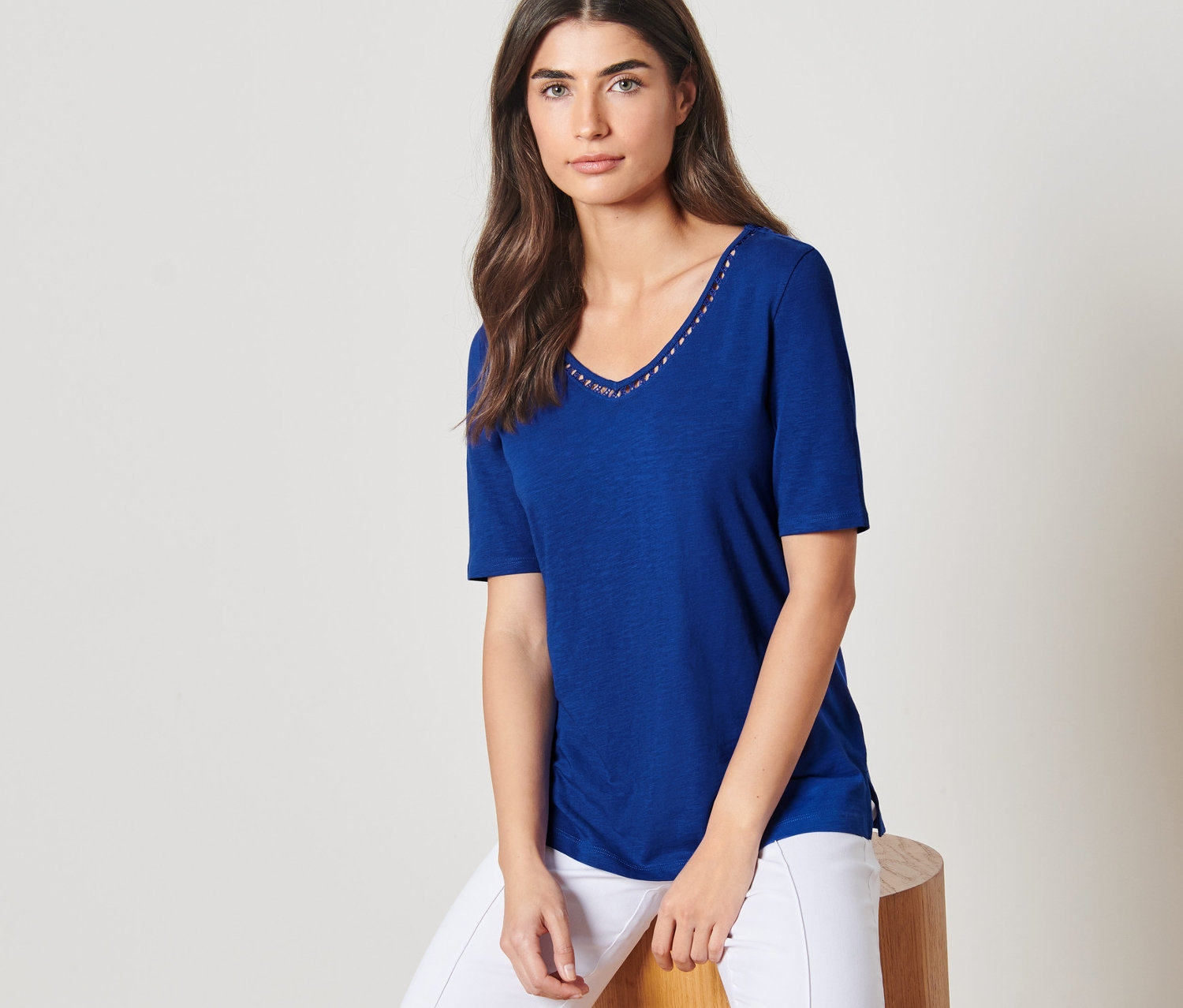 Frau sitzt auf einem Hocker und trägt ein kobaltblaues Shirt und eine weiße Hose.