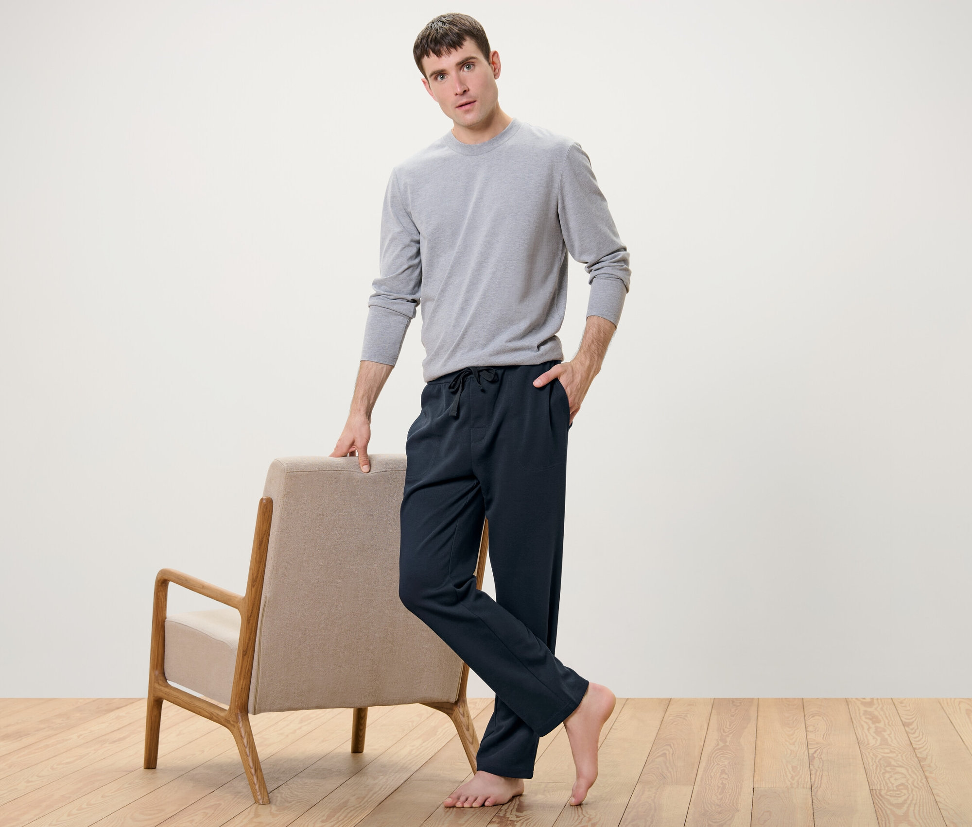 Mann posiert neben Sessel und trägt Homewearhose und graues Shirt.