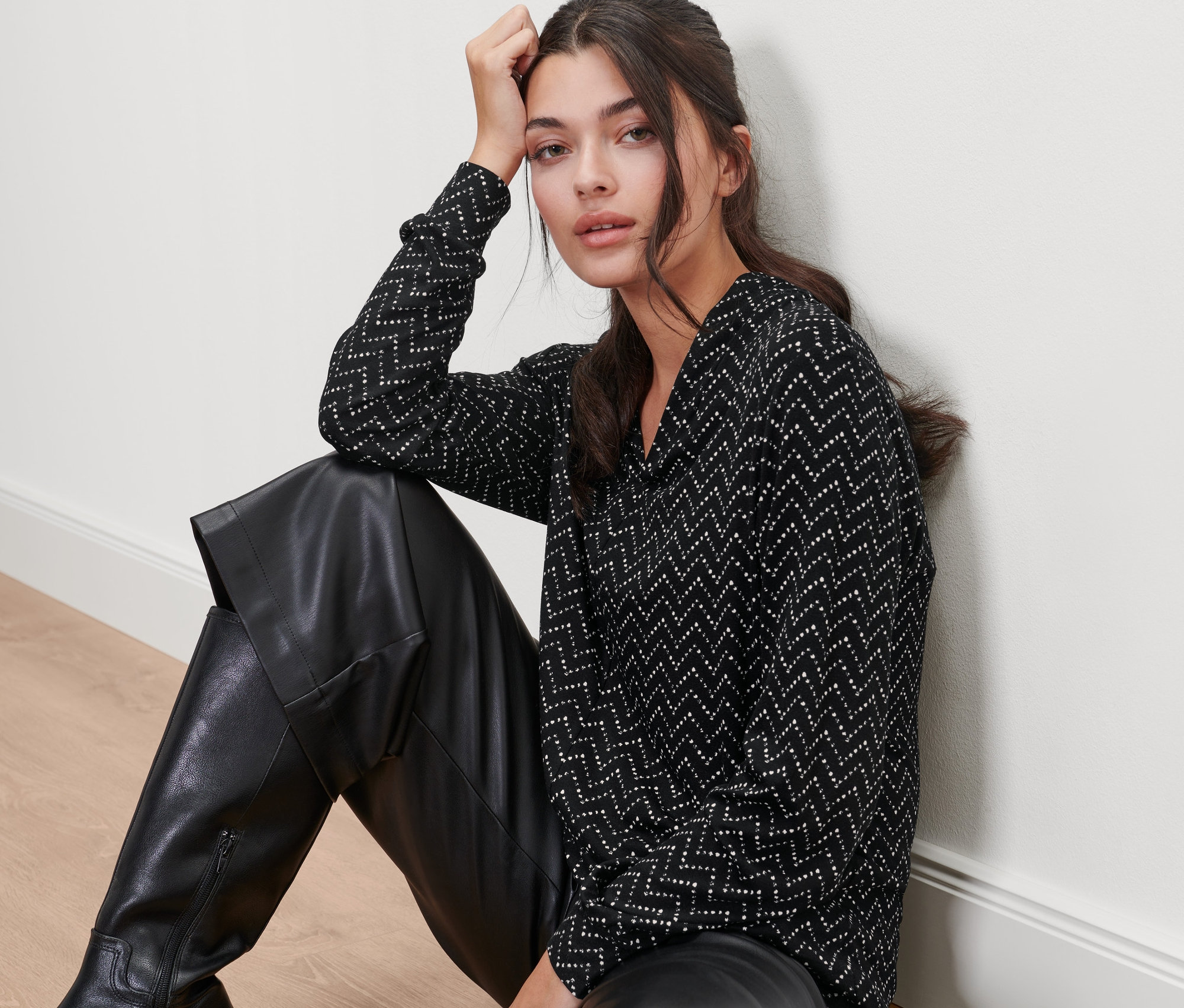 Eine Frau sitzt auf dem Boden und trägt eine schwarze Lederhose und ein Langarmshirt mit V-Ausschnitt, Alloverprint.