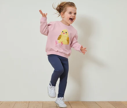 Mädchen springt in einem Kinder-Sweatshirt mit angerauter Innenseite und Leggings auf einem Holzboden.