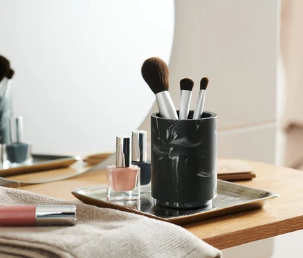 Make-up Pinsel in marmoriertem Utensilo neben Nagellacken auf einem Tablett.