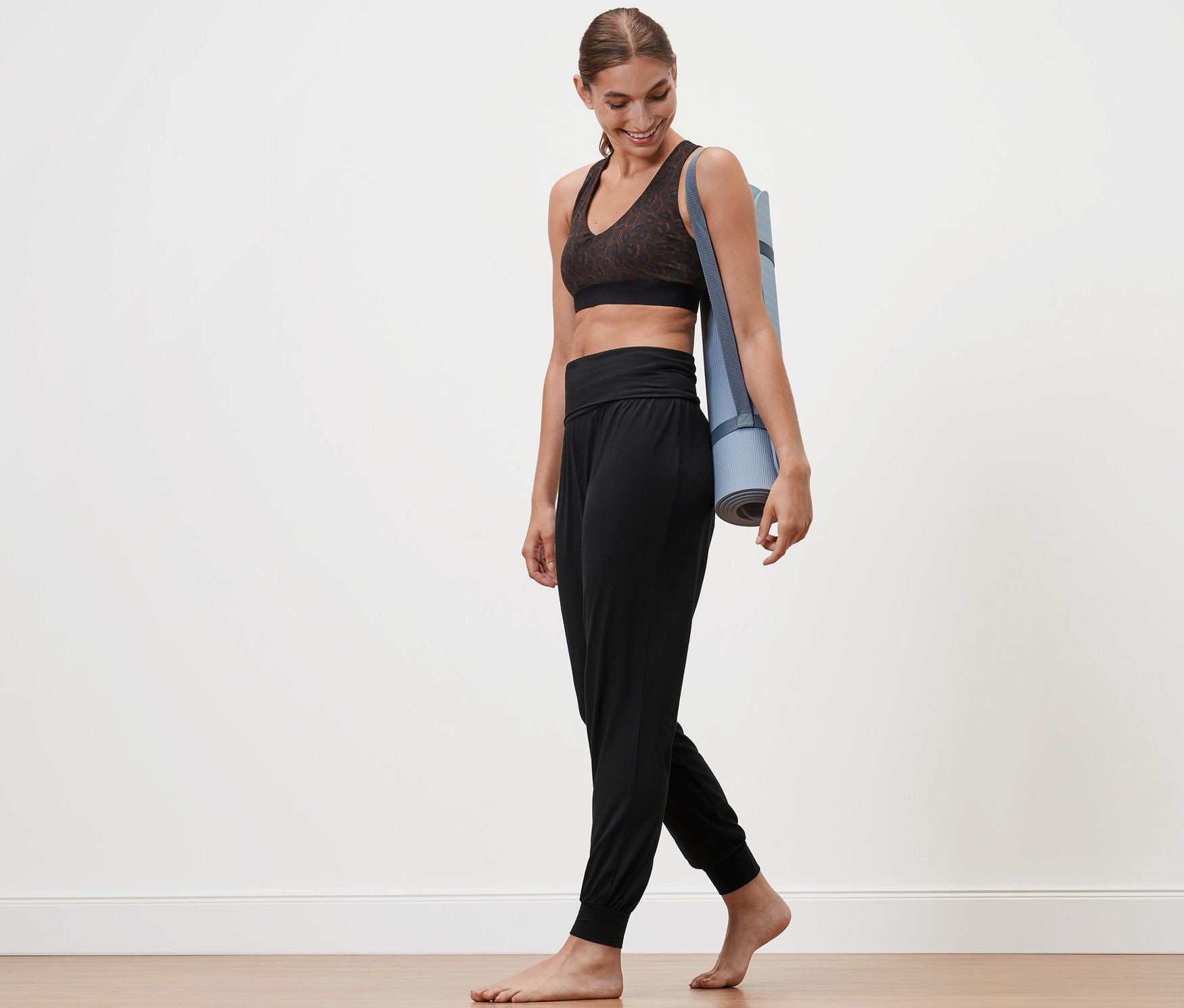Frau geht barfuß mit Yogamatte über der Schulter und trägt eine Yogahose.
