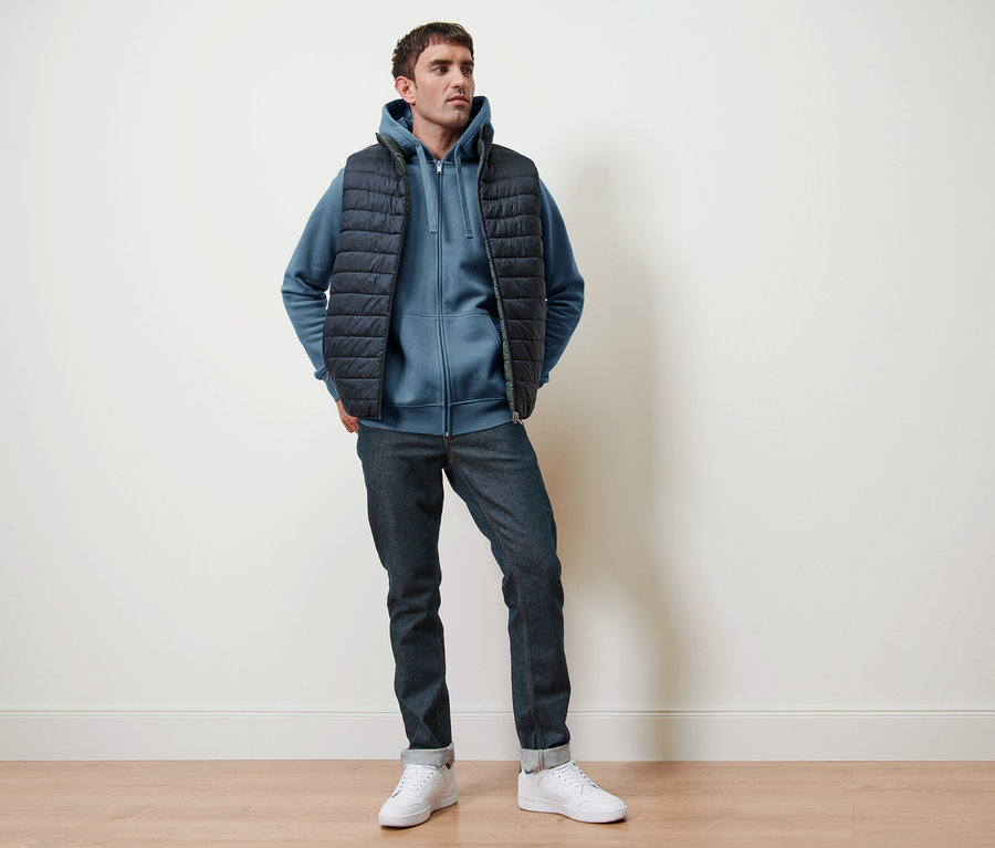 Mann posiert in rauchblauer Kapuzenjacke, Wendeweste und Jeans.