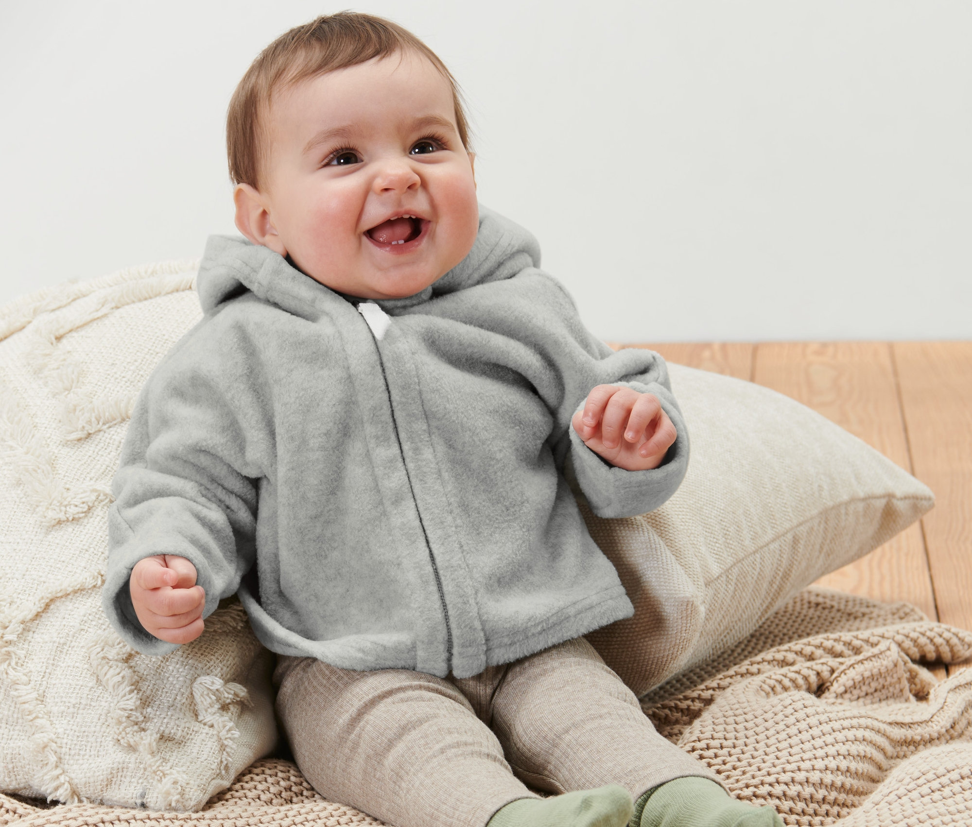 Lächelndes Baby sitzt auf einem Kissen und trägt eine graue Baby-Fleecejacke.