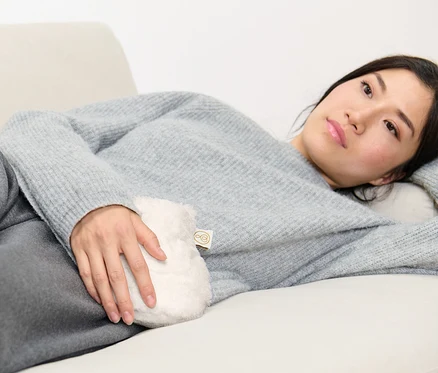 Frau liegt auf Couch und hält Mini-Wärmflasche.