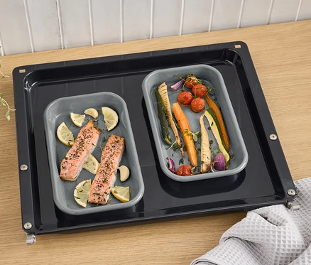 Zwei graue Emaille-Grillschalen mit Lachs, Zitrone und Gemüse auf einem Backblech.