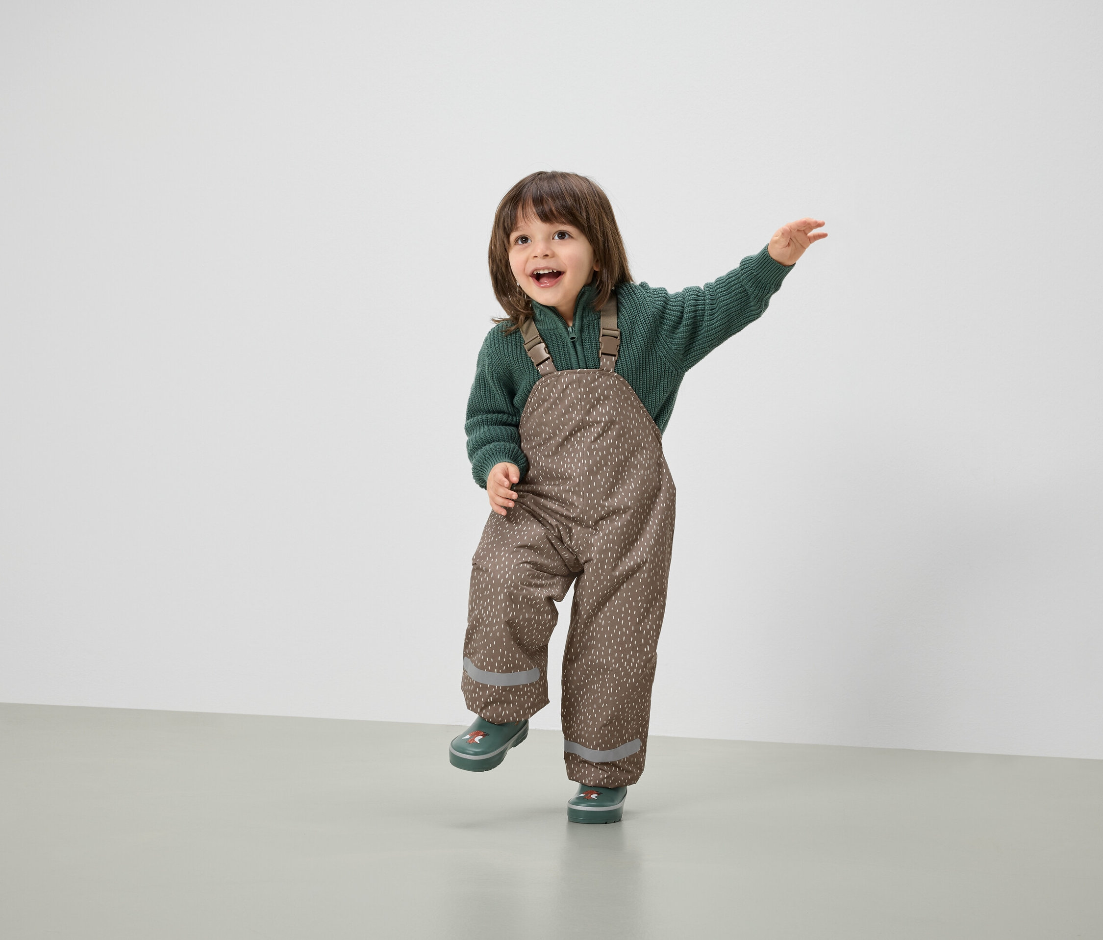 Ein lächelndes Kind posiert in braunen Latzhosen und grünem Pullover.