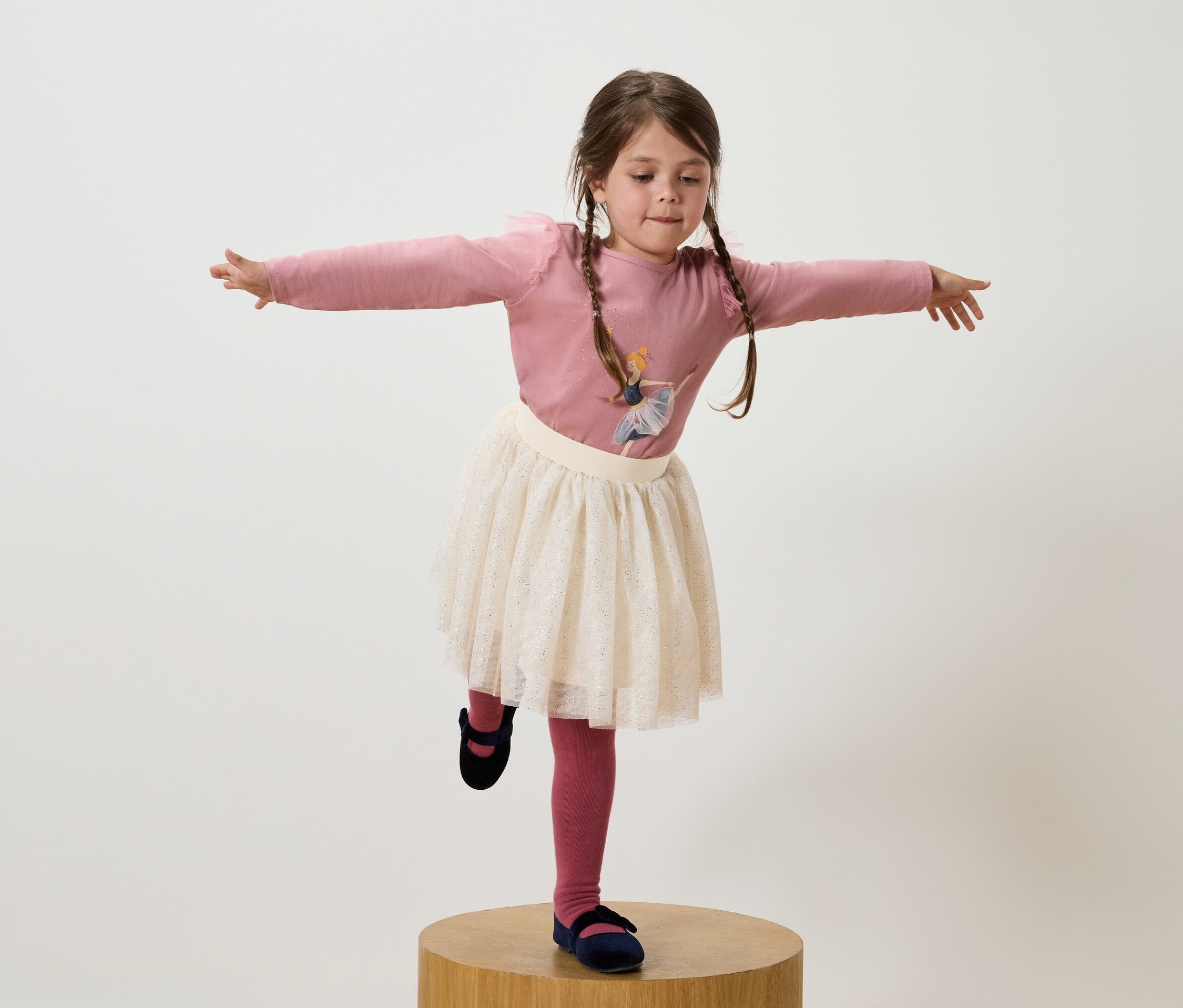 Mädchen mit braunen Zöpfen steht auf einem Bein auf einem Holzsockel, trägt ein rosa T-Shirt, einen weißen Rock und dunkelblaue Schuhe.