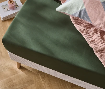Nahaufnahme eines Bettes mit olivgrünem Spannbettlaken, einer rosa Strickdecke und einem dekorativen Kissen.