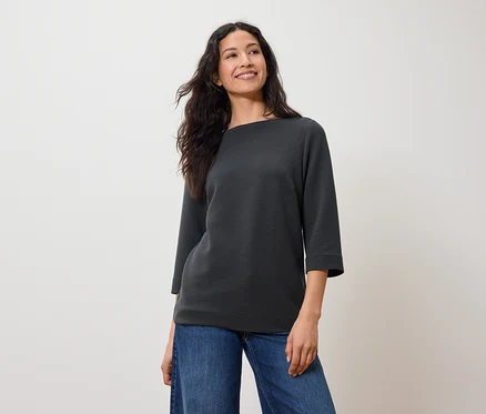 Frau mit dunklem Haar posiert in einem grauen Sweatshirt mit Dreiviertelärmeln und blauen Jeans.