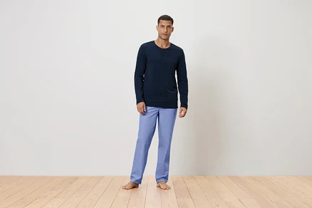 Mann steht barfuß auf einem Holzboden und trägt ein dunkelblaues Langarmshirt und hellblaue Hosen.