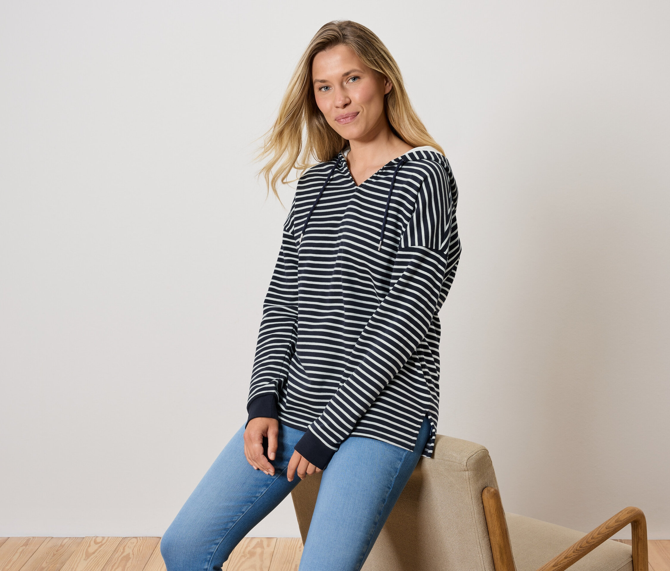 Eine Frau sitzt auf einem Stuhl, sie trägt blaue Jeans und ein gestreiftes Kapuzensweatshirt.