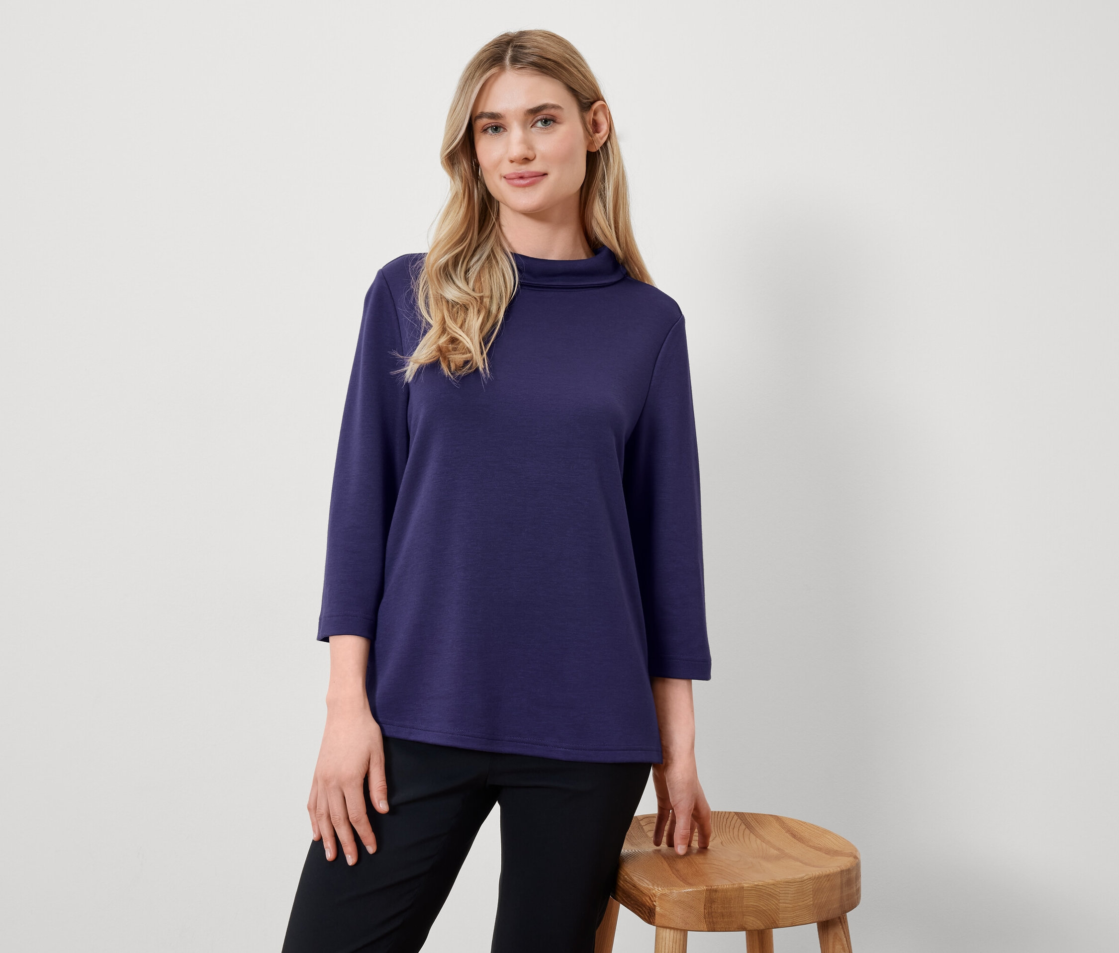Blonde Frau posiert in marineblauem Oberteil mit Dreiviertelärmeln und schwarzer Hose neben einem Holzhocker.