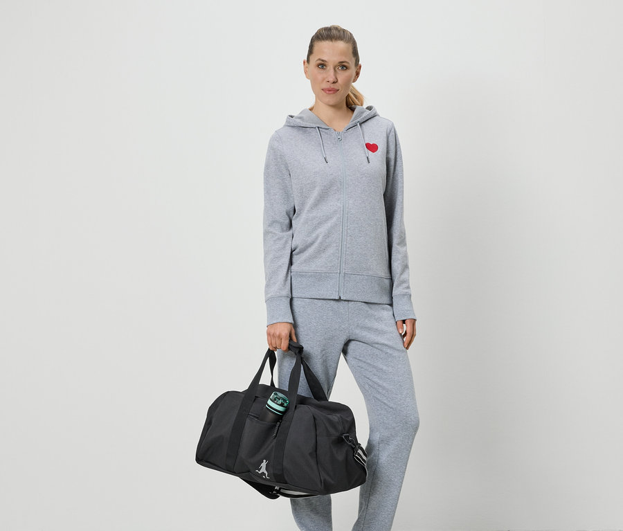 Frau in grauer Trainingsjacke mit rotem Herz und Jogginghose, die eine schwarze Sporttasche hält.