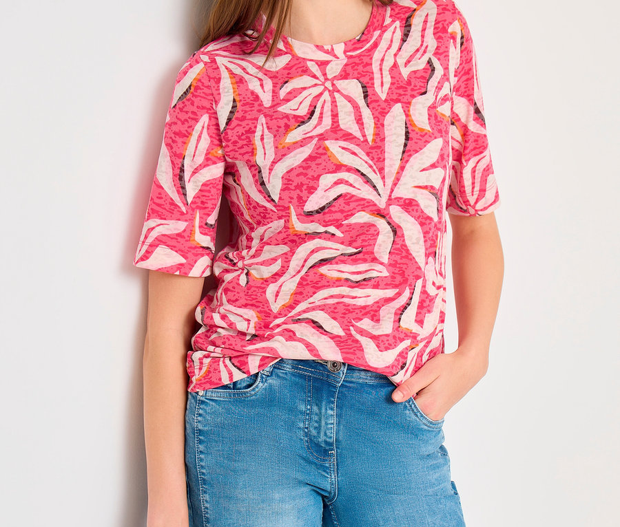 Frau trägt ein rosa CECIL Kurzarmshirt mit weißem Blattmuster und blaue Jeans, lehnt an einer weißen Wand mit einer Hand in der Hosentasche.
