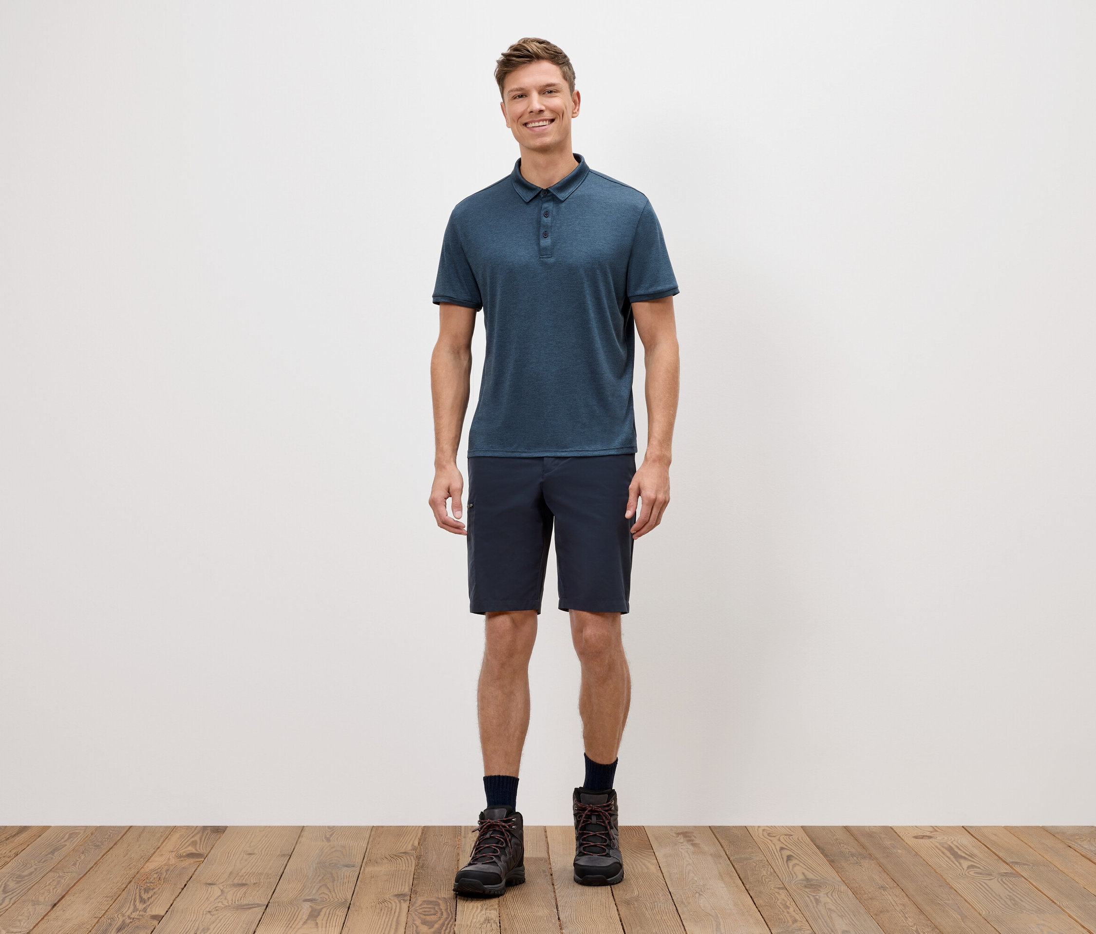 Ein Mann steht vor einer weißen Wand und trägt ein blaues Poloshirt, kurze Shorts und Wanderschuhe.