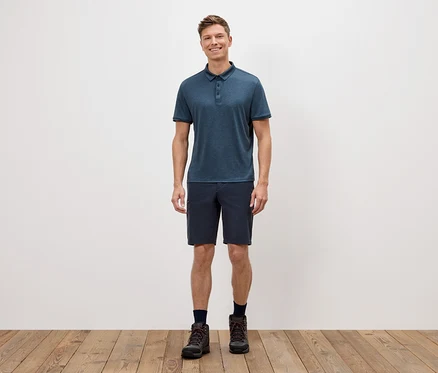 Ein Mann steht vor einer weißen Wand und trägt ein blaues Poloshirt, kurze Shorts und Wanderschuhe.