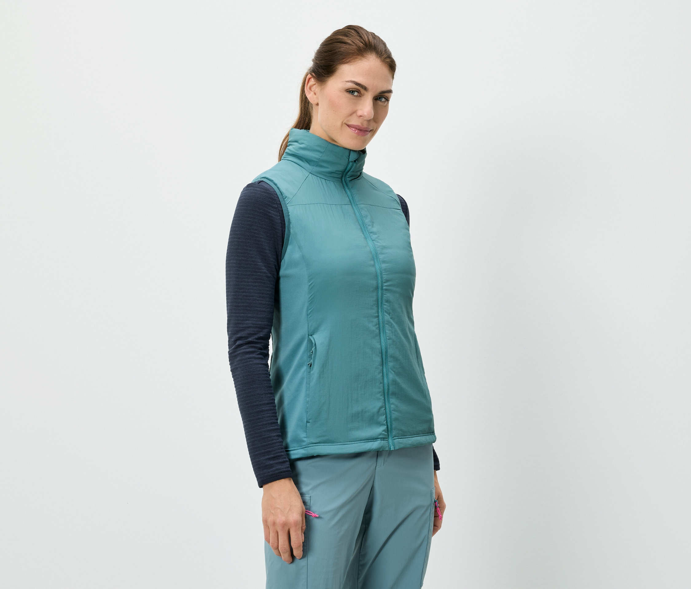 Eine Frau steht und trägt eine blaue Weste über einem langärmeligen Shirt, kombiniert mit blauen Hosen.