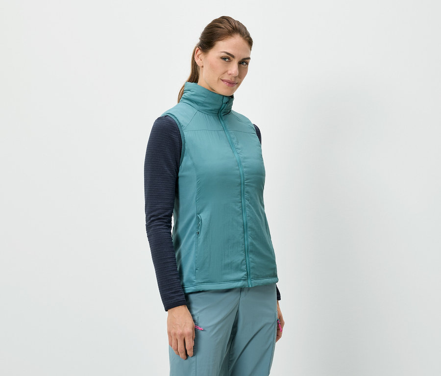 Eine Frau steht und trägt eine blaue Weste über einem langärmeligen Shirt, kombiniert mit blauen Hosen.