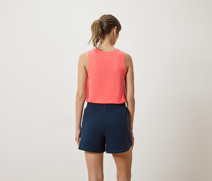 Rückenansicht einer Frau mit Pferdeschwanz, die ein korallfarbenes Tanktop und marineblaue Shorts vor einem cremefarbenen Hintergrund trägt.