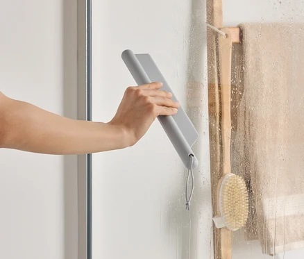 Eine Hand wischt mit einem grauen Abzieher eine Duschwand ab, neben einer Holzleiter mit einem Handtuch und einer Bürste.