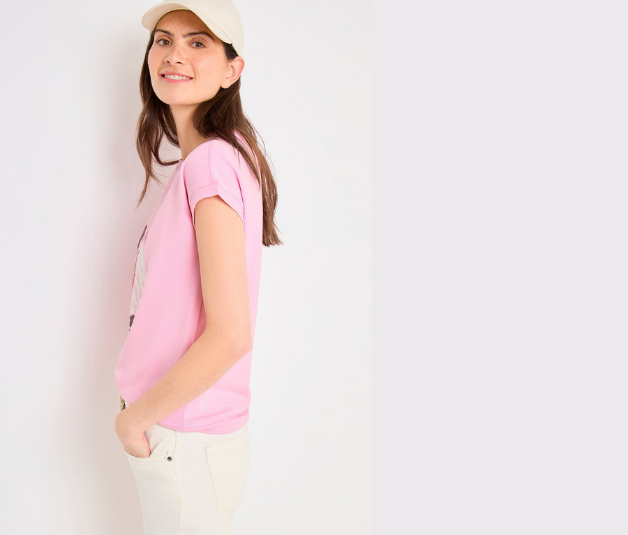 Lächelnde Frau in beiger Kappe und heller Hose, trägt ein pinkes CECIL Placement-Shirt mit hochgekrempelten Ärmeln, Hände in den Taschen.