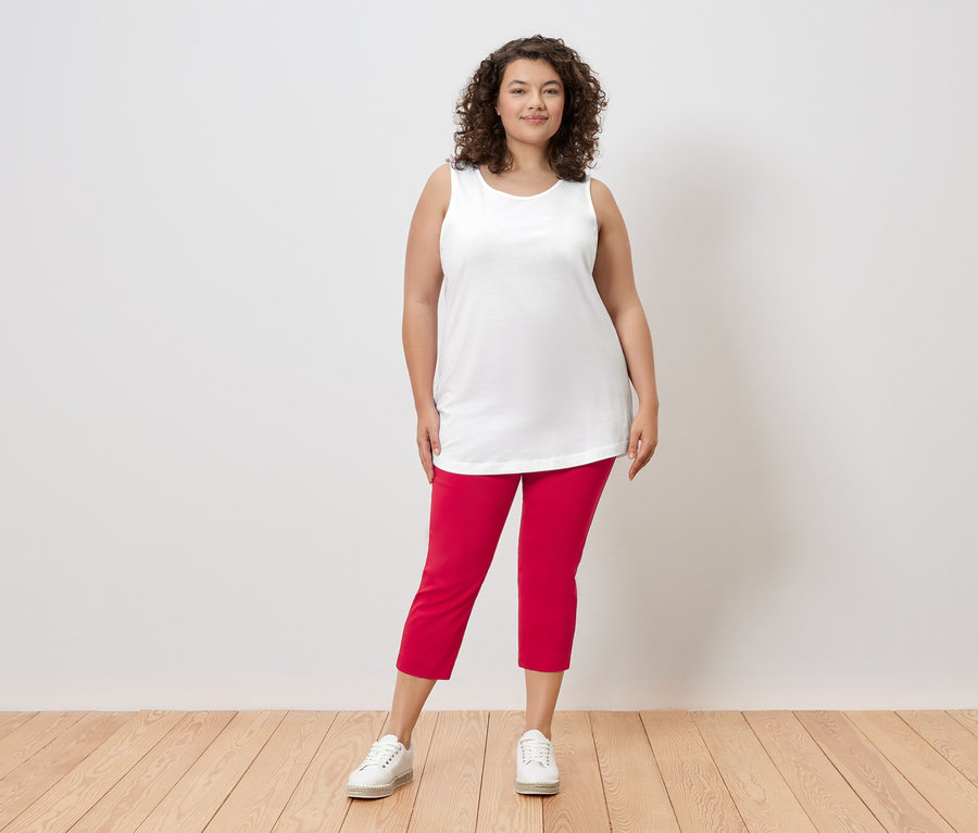 Eine Frau mit lockigem Haar steht vor einer Wand und trägt ein weißes Top, rote Hosen und weiße Turnschuhe.