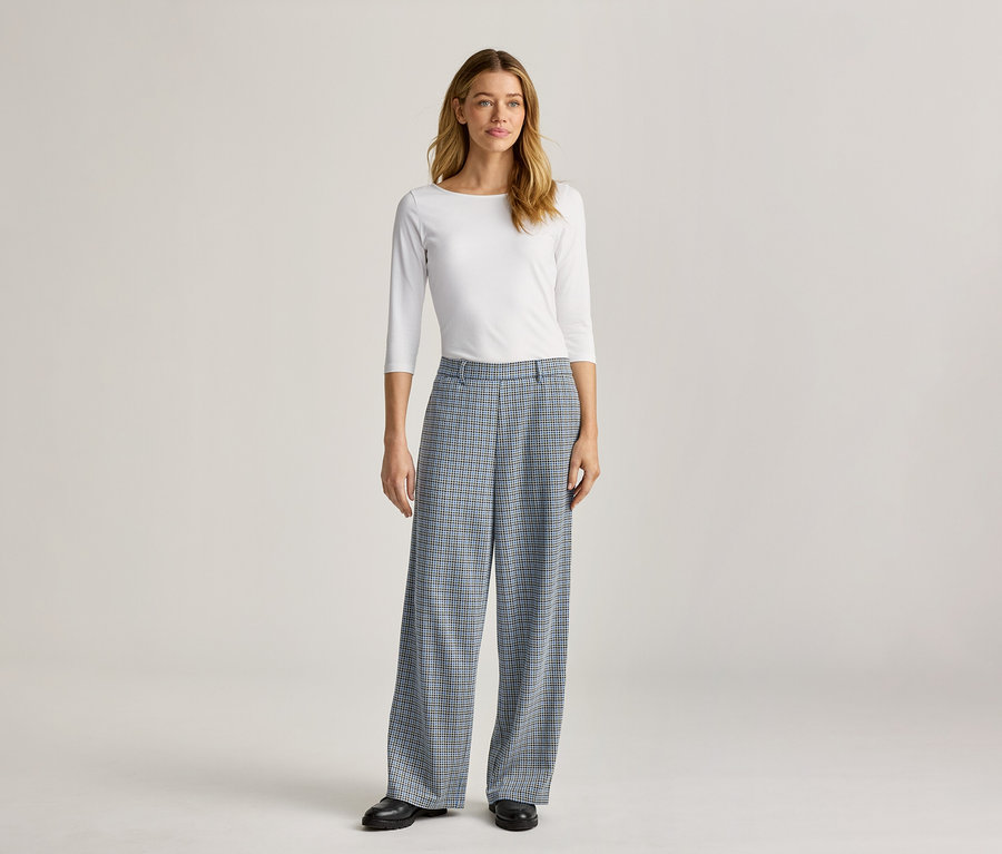 Eine Frau steht in einem weißen, langärmeligen Oberteil und einer weiten, blau karierten Hose mit schwarzen Schuhen.