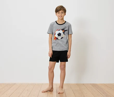 Ein Junge steht barfuß vor einer weißen Wand und trägt ein graues T-Shirt mit einem Fußballaufdruck und schwarzen Shorts.