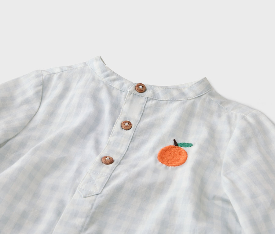 Nahaufnahme eines hellblau-weiß karierten Hemdes mit Mandarin-Kragen, Knopfleiste mit drei Knöpfen und einer aufgestickten Orange.