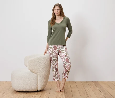 Eine Frau steht neben einem Sessel und trägt einen olivgrünen Pullover und eine geblümte Hose.