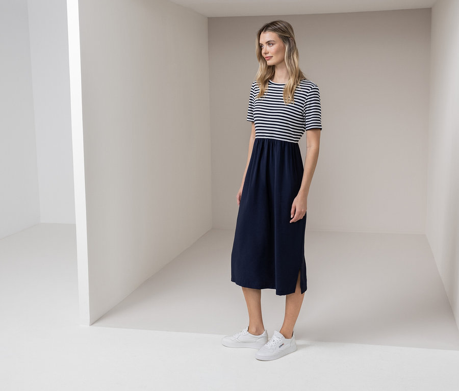 Frau steht in einem weißen Raum und trägt ein blau-weiß gestreiftes Kleid und weiße Turnschuhe.