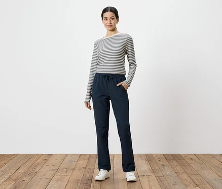 Frau mit grau-weiß gestreiftem Langarmshirt und marineblauer Hose mit Händen in den Taschen, vor weißem Hintergrund.