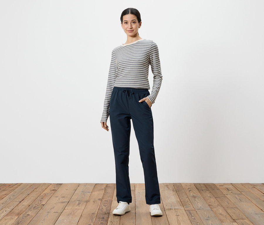 Frau mit grau-weiß gestreiftem Langarmshirt und marineblauer Hose mit Händen in den Taschen, vor weißem Hintergrund.