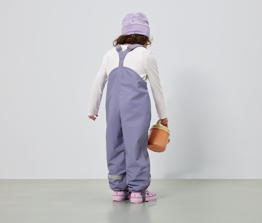 Rückansicht eines Kindes mit lila Mütze und Overall, weißem Langarmshirt, rosa Schuhen und einem orangefarbenen Eimer.