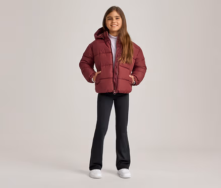 Lächelndes Mädchen in bordeauxroter Steppjacke mit Kapuze, schwarzer Schlaghose und weißen Turnschuhen, die Hände in den Jackentaschen.