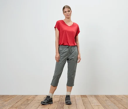 Frau steht in rotem T-Shirt und grauen Dreiviertelhosen vor einer weißen Wand.