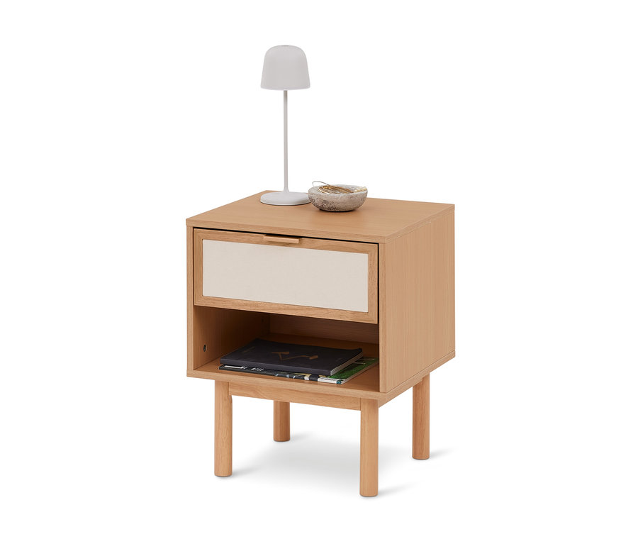 Nachttisch aus hellem Holz mit einer Schublade und einem offenen Fach. Darauf stehen eine weiße Lampe und eine kleine Schale, im Fach liegen zwei Bücher.