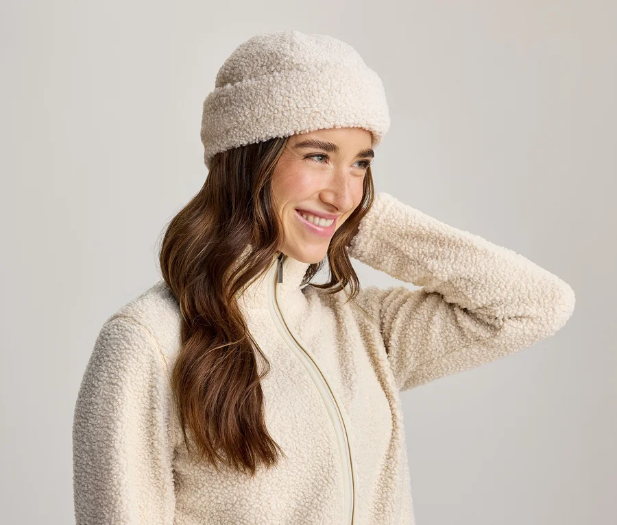 Lächelnde Frau mit langen braunen Haaren, die eine cremefarbene Teddyfell-Mütze und eine passende Reißverschlussjacke trägt, ihre Hand am Hinterkopf.