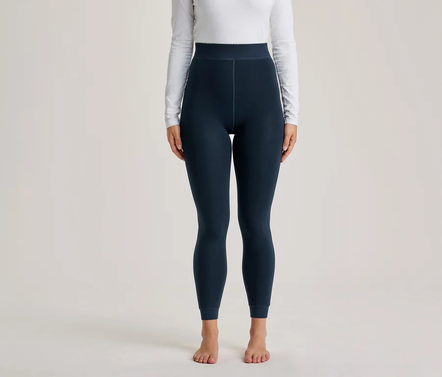 Eine Person trägt ein weißes Langarmshirt und dunkelblaue Leggings, steht barfuß vor einem schlichten Hintergrund.