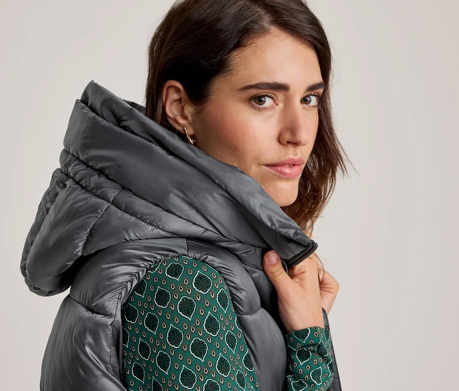 Nahaufnahme einer Frau in einer grauen, gesteppten Kapuzenjacke über einem grün gemusterten Hemd, die direkt in die Kamera blickt.