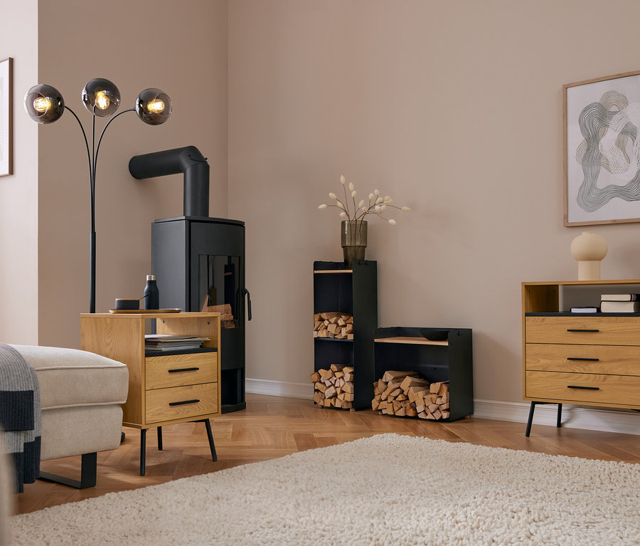 Modernes Wohnzimmer mit schwarzem Kaminofen, einem hohen Kaminholzregal mit Ablagefläche und einem breiten Kaminholzregal mit Ablagefläche, beide gefüllt mit Brennholz. Ein helles Sofa, ein Holztisch und eine Kommode stehen auf einem Teppich.