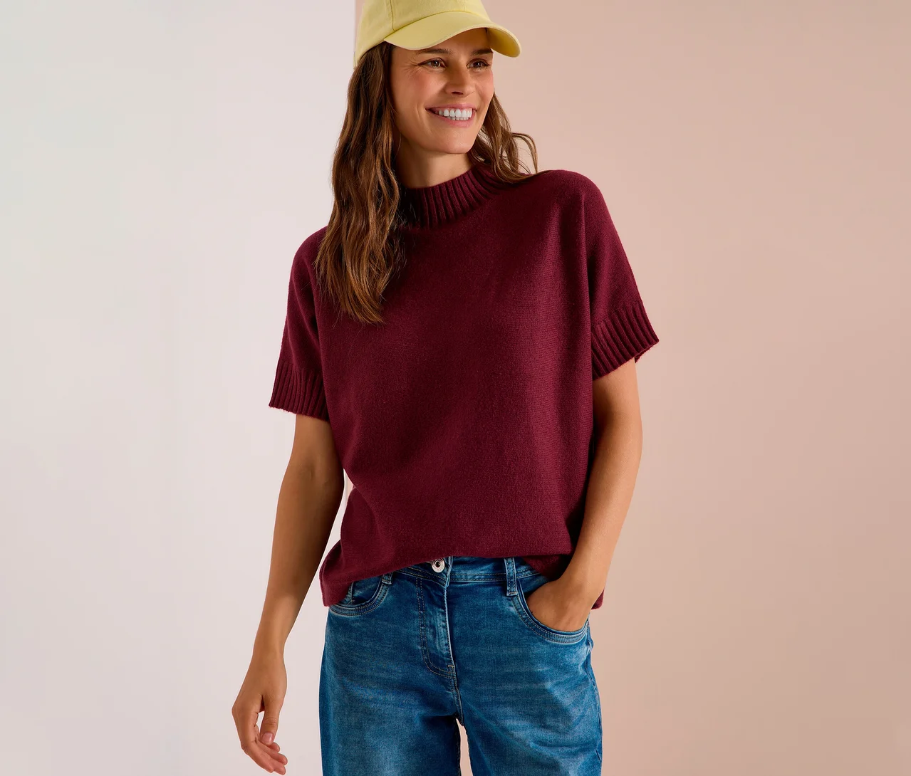 Lächelnde Frau mit gelber Kappe, die einen bordeauxroten CECIL Strickpullover und Jeans trägt.