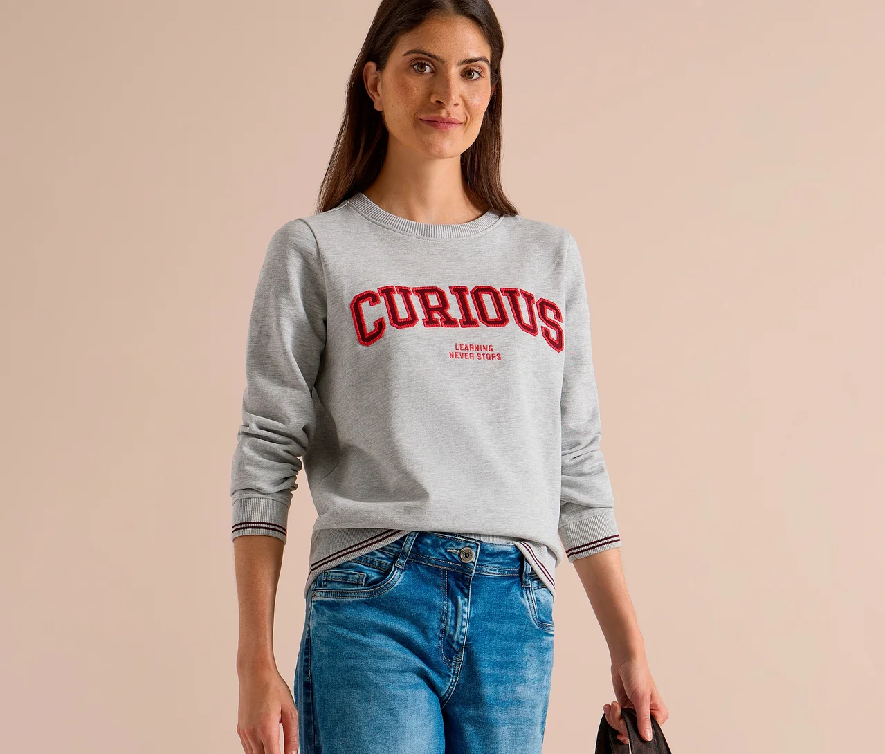 Eine Frau trägt ein graues CECIL Sweatshirt mit der Aufschrift „CURIOUS LEARNING NEVER STOPS“ und blaue Jeans.