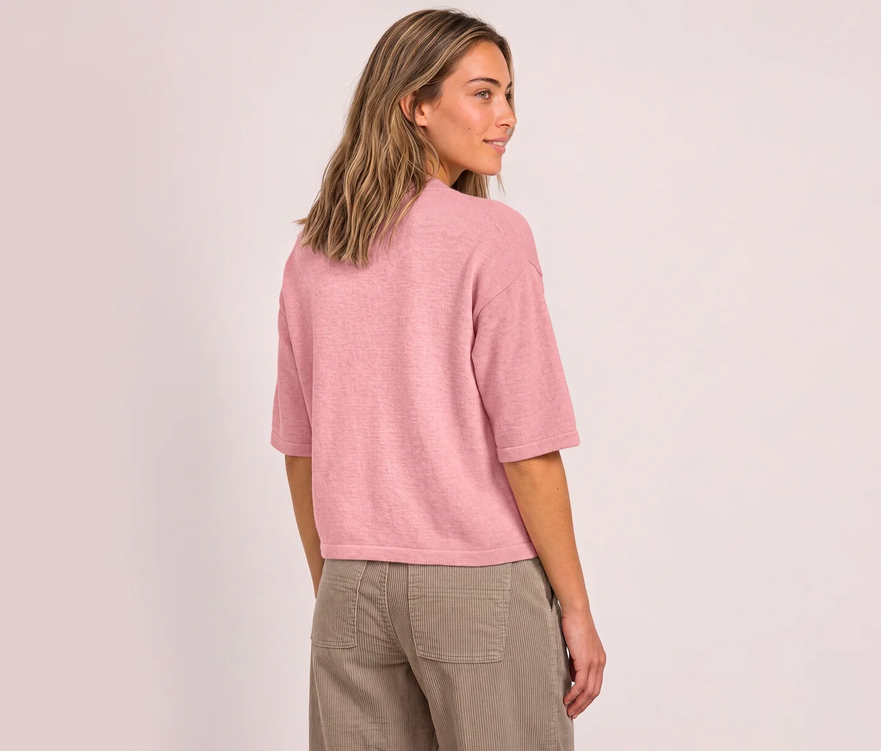 Eine Frau mit langen, hellbraunen Haaren, von hinten gesehen, blickt über ihre rechte Schulter. Sie trägt einen pinken Street One 3/4-Strickpullover und eine hellbraune Cordhose.