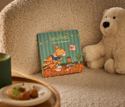 Ein beiges, flauschiges Kissen mit dem Buch »Leo Lausemaus & der Wichtelbesuch« und einem hellbeigen Teddybär. Links davor steht eine brennende grüne Kerze und ein Teller mit Keksen.
