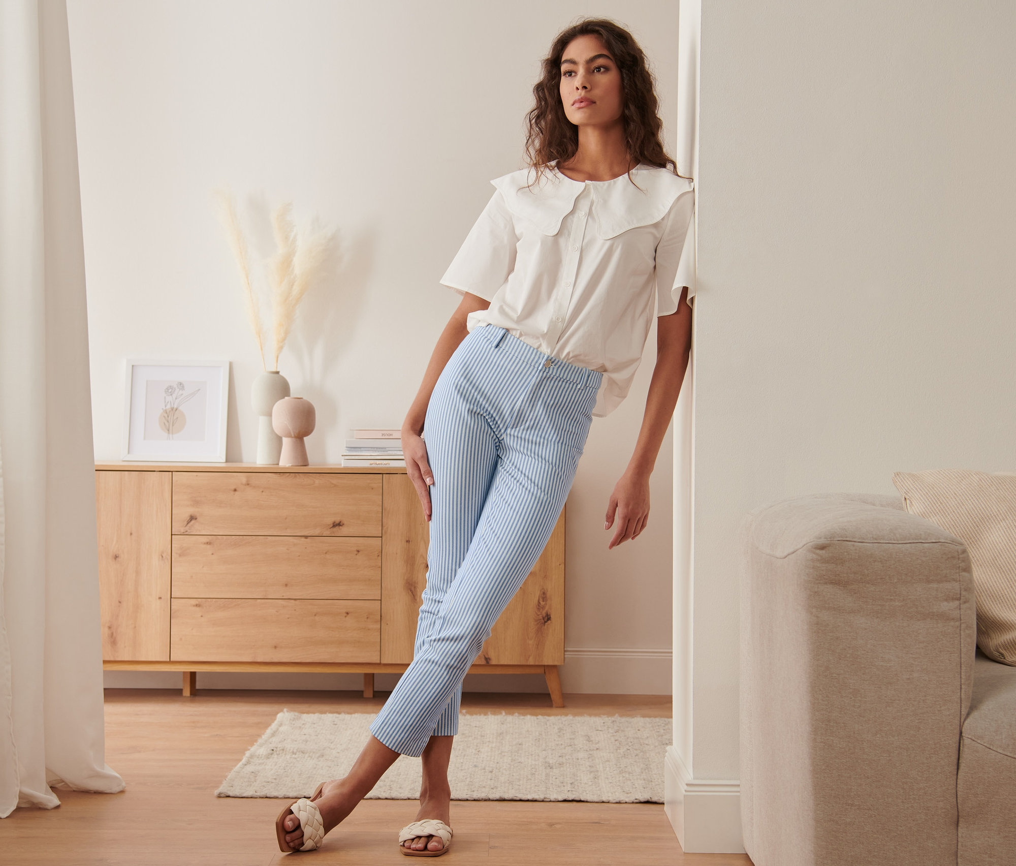 Frau lehnt an Wand, trägt gestreifte Stretchhose und weiße Bluse.