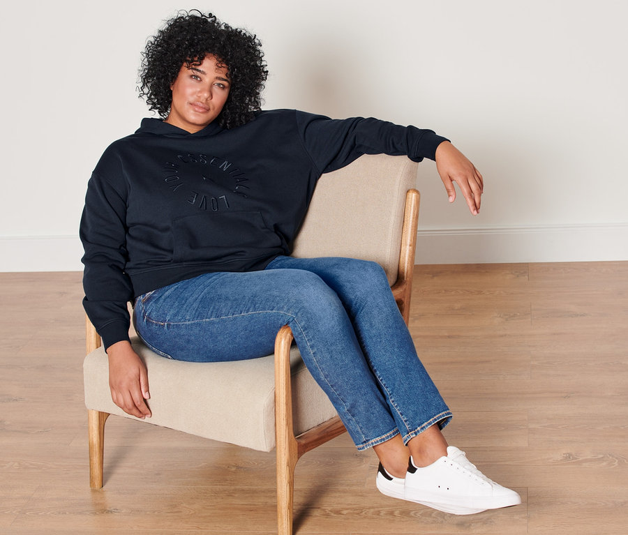 Frau sitzt in Sessel, trägt dunkelblauen Hoodie und Slim-Denim »Fit Emma« in Mid Blue.