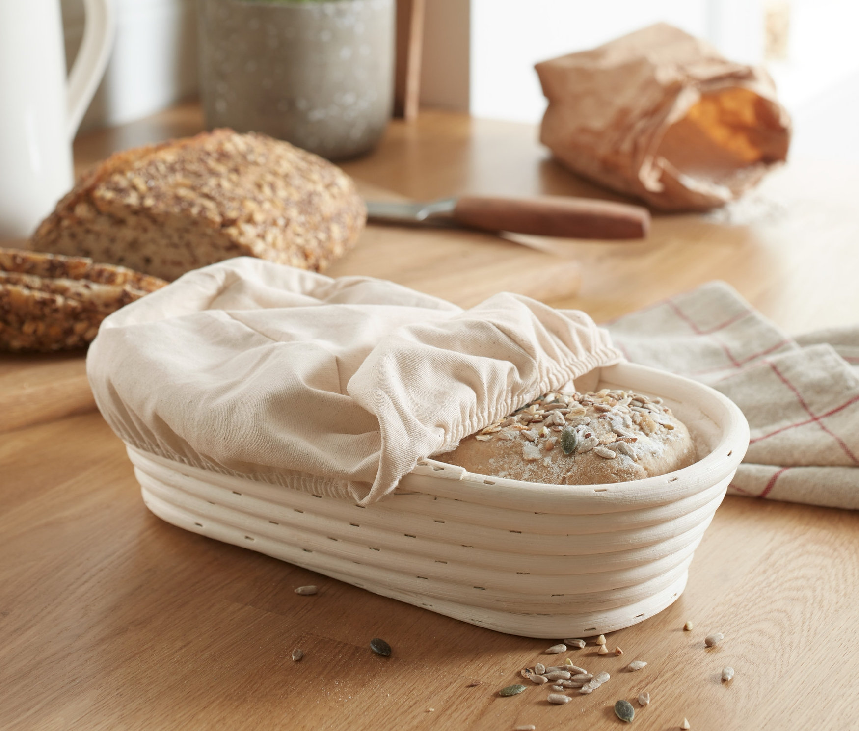 Brot in einem Gärkörbchen, oval, mit einem Tuch bedeckt, liegt auf einer Holzplatte.