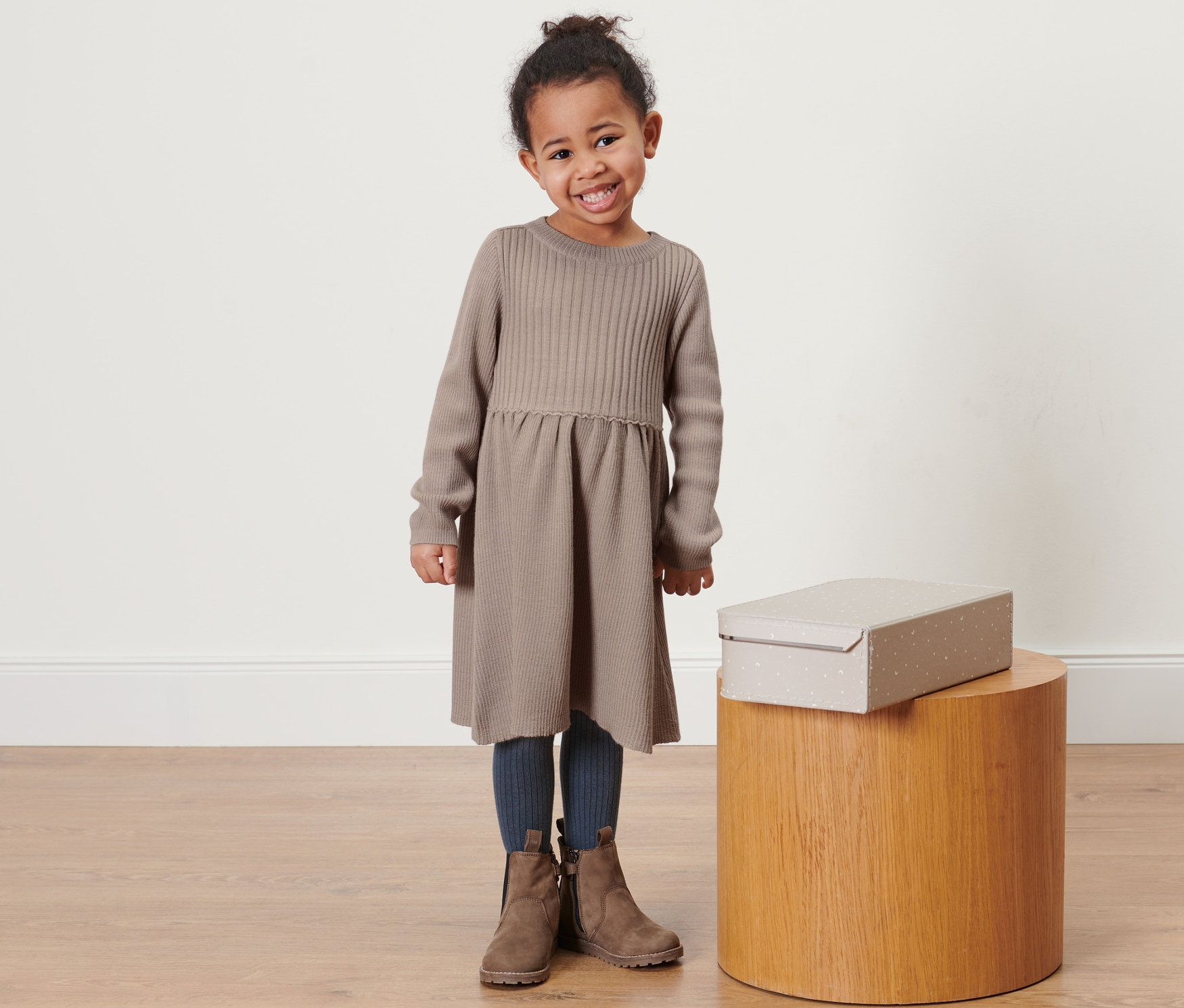 Ein lächelndes kleines Mädchen posiert in einem hellbraunen Kleinkind-Merino-Strickkleid. Eine Schachtel steht auf einem Holzhocker neben ihr.